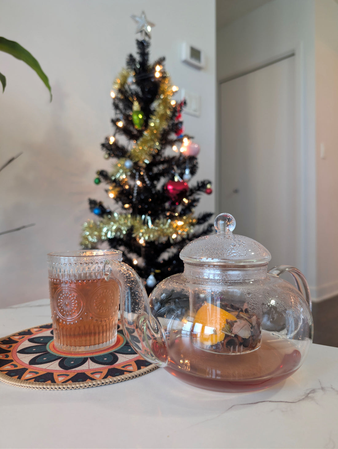 Glass tea kettle and glass cup with Christmas themed hibiscus tea in front of a Christmas tree